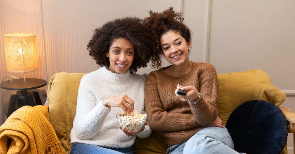 Duas mulheres sentadas no sofá assistindo filmes e séries pela TV, com pipoca e controle remoto na mão.