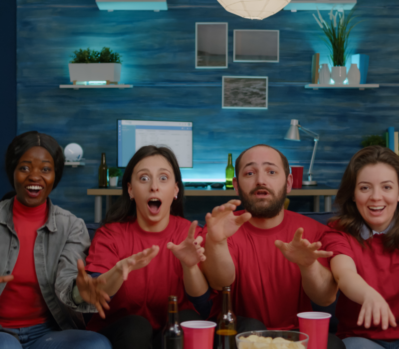 Grupo de amigos reagindo com emoção e surpresa ao assistir TV juntos em um ambiente descontraído.